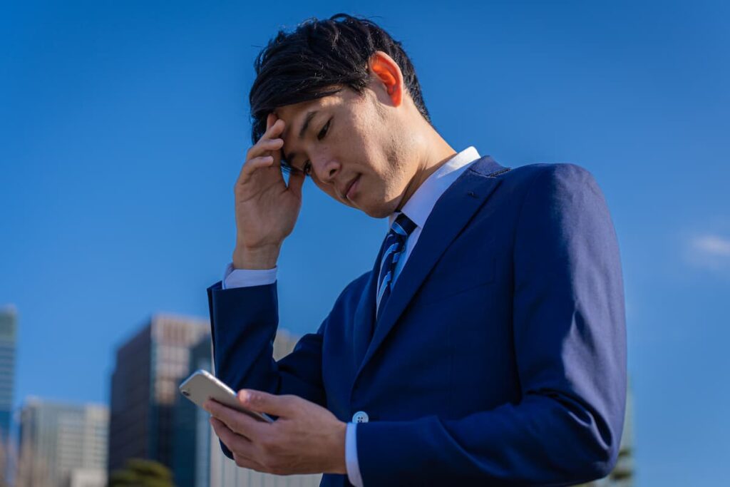 屋外でスマートフォンを見ながら悩むスーツ姿の男性
